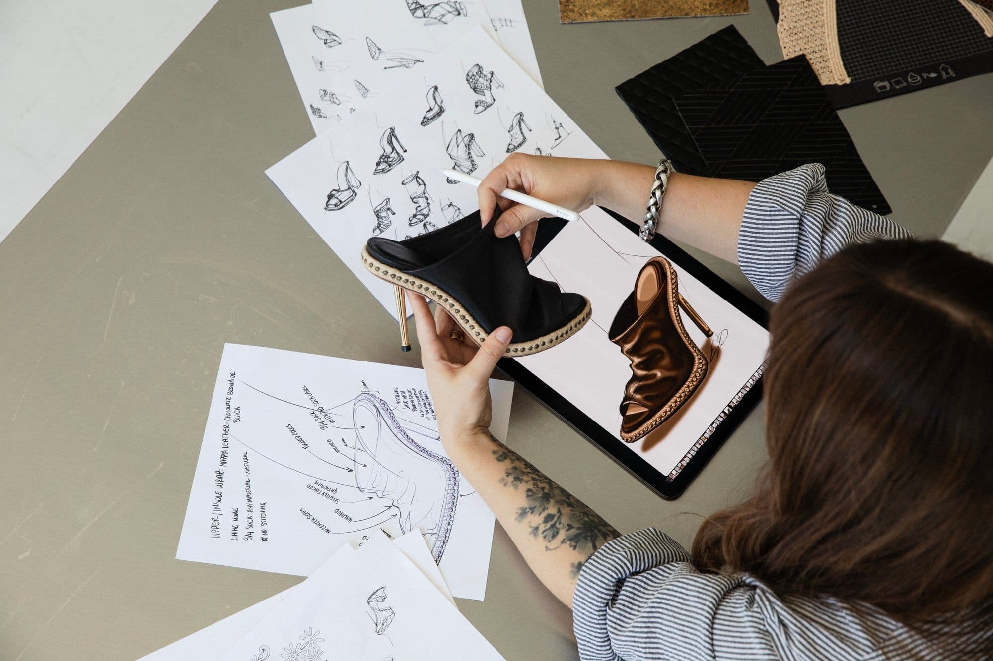 Erin Lavigne holding a black leather mule sample beside its digital rendering, surrounded by sketches and material swatches